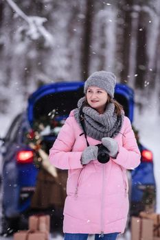 A woman in a winter snow-covered forest in the trunk of a car decorated with Christmas decor. A female photographer holds a camera in her hands.