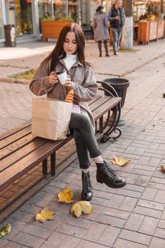 A stylish brunette woman walks around the autumn city. The brunette is sitting on a bench outside with a glass of coffee and a phone, holding a craft bag with a baguette.