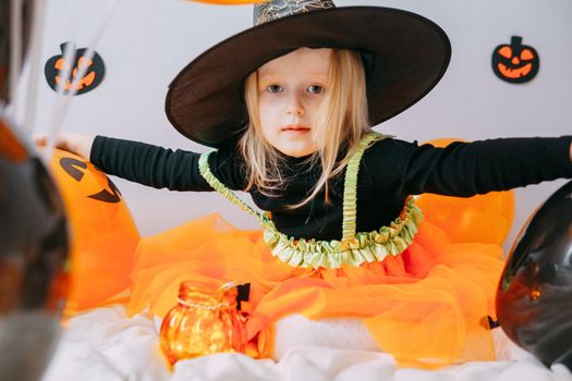 Children's Halloween - a girl in a witch hat and a carnival costume with airy orange and black balloons at home. Ready to celebrate Halloween.