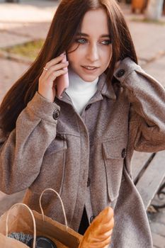 A stylish brunette woman walks around the autumn city. The brunette is sitting on a bench outside with a glass of coffee and a phone, holding a craft bag with a baguette.