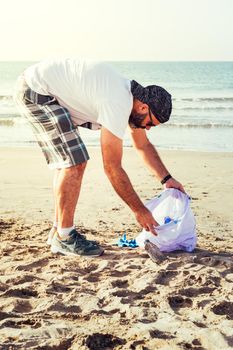 vertical photo of an activist picking up plastics and trash that pollutes the sea on a bag on a sunny day, cleaning on the beach, campaign to clean volunteer concept