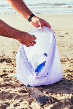 vertical photo of the hands of an activist picking up plastics and trash that pollutes the sea on a bag on a sunny day, cleaning on the beach, campaign to clean volunteer concept
