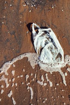 vertical photo of a plastic bag on the beach, seashore and water pollution with plastics concept, waste at the seaside, top view of an environmental problem with wave of water and sand