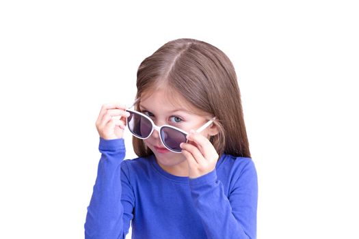 Smiling child holding white sunglasses down on eyes and look out, isolated on white background looking at camera waist up caucasian little girl of 5 years in blue