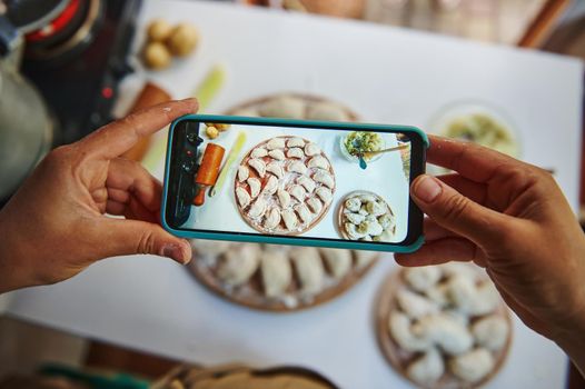 Selective focus on the hands of a food blogger, holding smartphone in live view mode, and taking photo of dumplings on the wooden board, for the blogging content. Ukrainian national dish Varenyky