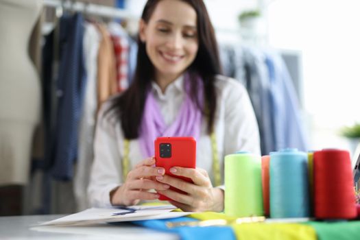 Beautiful fashion designer is sitting at table and typing on smartphone. Workshop full of colorful details of fabrics and sewing supplies