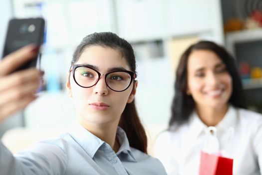 Young woman in glasses holds phone in front of face and smiles at colleagues in office. Video call or selfie at work