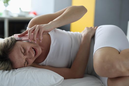 Sick woman is lying in bed and talking on smartphone with doctor. Stomach problems suffering from abdominal pain