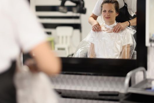 Female hairdresser puts paper collar bag around woman neck to protect clothes she cuts hair indoors. Caucasian beautiful lady changes image in a hairdresser