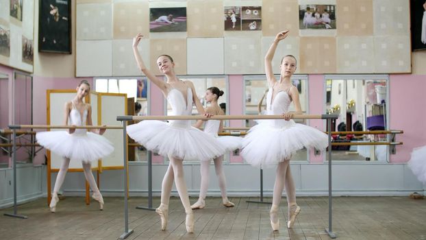 in the ballet hall, girls in white ballet tutus, packs are engaged at ballet, rehearse croise forward, Young ballerinas standing on toes in pointe shoes at railing in ballet hall. High quality photo