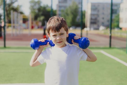 A child with dumbbells on the playground. A sporty kid. Illustrating an article about sports. Children's exercises. Education of champions. Light weightlifting. Lifestyle