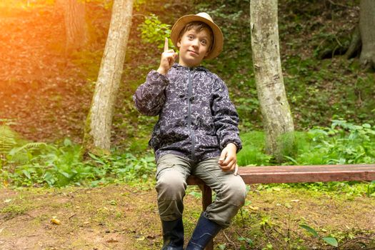 funny Caucasian boy in a traveler's hat and windbreaker is sitting on a bench in a nature park near a forest stream and rocks, looking at the camera and raised his index finger up