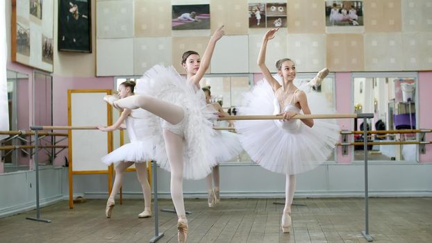 in ballet hall, girls in white ballet skirts are engaged at ballet, rehearse attitude, Young ballerinas standing on toes in pointe shoes, raise legs up behind elegantly, at railing in ballet hall. High quality photo