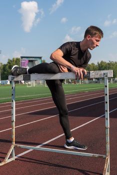 Young sporty man athlete runner in sportswear stretching before running hurdles on stadium