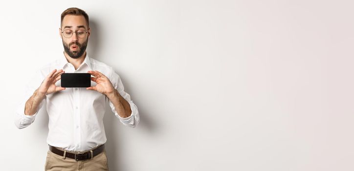 Amused handsome guy showing mobile screen, looking excited at online website, standing over white background.