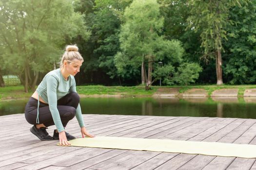 A woman in sportswear, in the summer by pond, unwinds a green gym mat, prepares for sports. Copy space