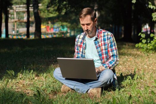 Adult man works on his computer in the park on the lawn.Remote work concept. The writer works remotely, enjoying nature.