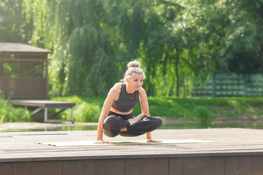 A beautiful woman in a gray top and leggings, sitting on a wooden platform by a pond in a park in summer, does yoga with her legs crossed and holding on to her hands. Copy space