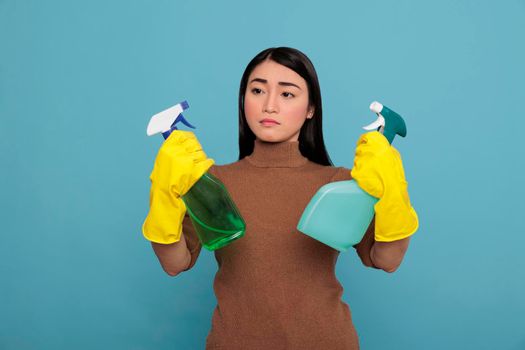 Asian tired housewife frustrated from overwork holding two detergent sprayers in yellow gloves isolated on a blue background, Housekeeping and hygiene concept, Daily homework