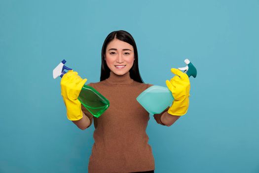 Cheerful joyful asian woman holding two detergent spray in yellow gloves isolated on a blue background, Cleaning home concept, smiling happy housewife worker female at chores