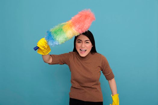 Angry energetic young asian houseworker holding colorful dust brush in yellow glove isolated against a blue background, Cleaning home concept, Crying female with face expression