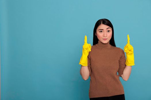 Humble confidence asian houseworker wearing yellow gloves and pointing fingers upward to copy space Isolated on a blue background, Cleaning home concept, faithful positive housekeeper