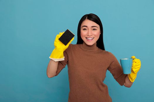 Excited smiling asian young housemaid wearing yellow gloves holding a sponge and coffee cup Cleaning home concept, Refreshment tea at break time, Woman feeling cheerful satisfied and positive