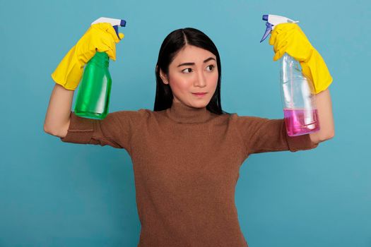 Strong serious asian housemaid raising arms from chores holding two detergent sprays in the yellow gloves isolated on a blue background, Cleaning home concept, Energetic confidence woman at work