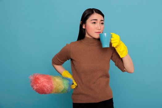 Sad upset and depressed of young houseworker taking coffee in the yellow glove with colorful duster brush on other hand isolated on a blue background, Cleaning home, Refreshment tea break time