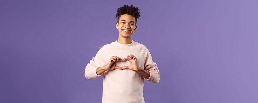 Portrait of young happy, romantic lovely boyfriend express his love and sympathy to dearest person, show heart sign and beaming smile, like this girl and trying to confess feelings, purple background.