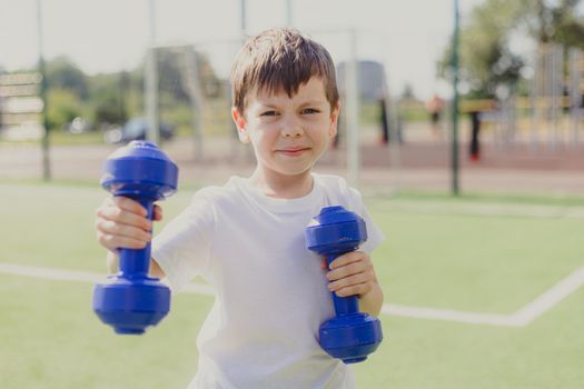 A child with dumbbells on the playground. A sporty kid. Illustrating an article about sports. Children's exercises. Education of champions. Light weightlifting. Lifestyle