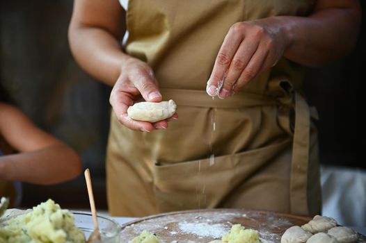 Details: hands of a housewife in chef's apron, sprinkling white flour on a wooden board while hands-on dumplings, cooking homemade varenyky in the country kitchen. Scene of artisanal food preparation