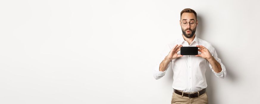 Amused handsome guy showing mobile screen, looking excited at online website, standing over white background.