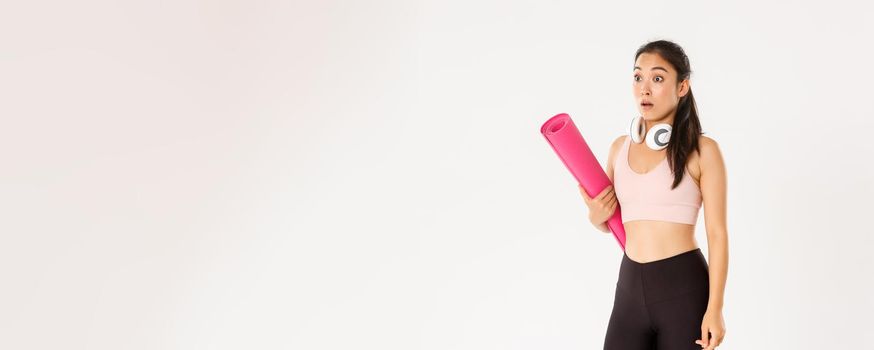 Sport, wellbeing and active lifestyle concept. Shocked asian fitness girl in stupor, drop jaw and gasping while looking left at logo or banner info, holding rubber mat for fitness exercises.