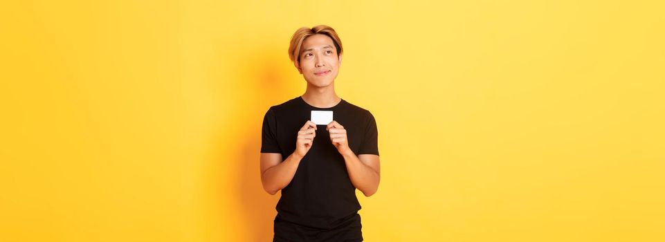 Thoughtful smiling asian guy thinking while showing credit card, looking upper left corner dreamy, yellow background.