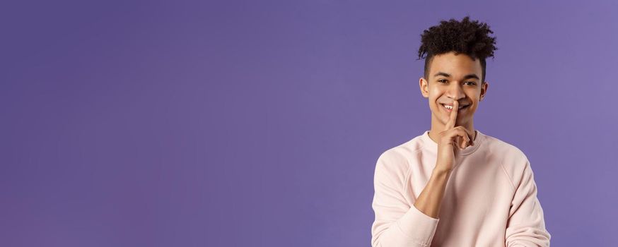 Close-up portrait of attractive smiling young man hiding secret, asking keep silent or quiet to do surprise, show shush gesture, place index finger to mouth, standing purple background.