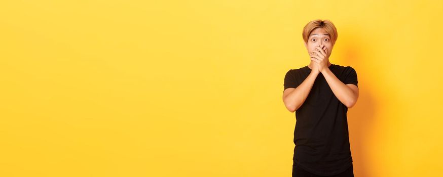 Portrait of surprised gasping asian guy with blond haircut, cover mouth with hands and looking amazed, standing yellow background.