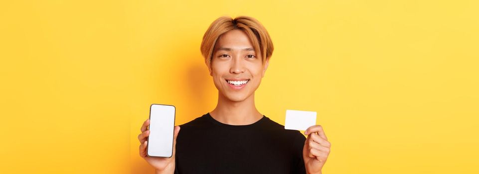 Close-up of smiling handsome asian guy showing smartphone screen and credit card, standing yellow background.