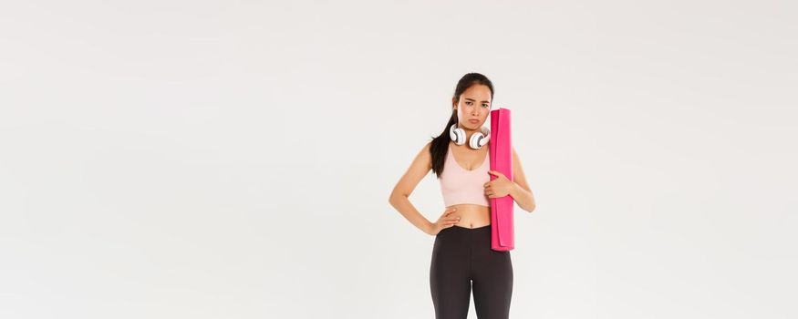 Full length of gloomy and upset cute brunette asian fitness girl in sportswear, sulking while holding rubber mat for yoga classes, looking disappointed as gym closed during covid-19 quarantine.