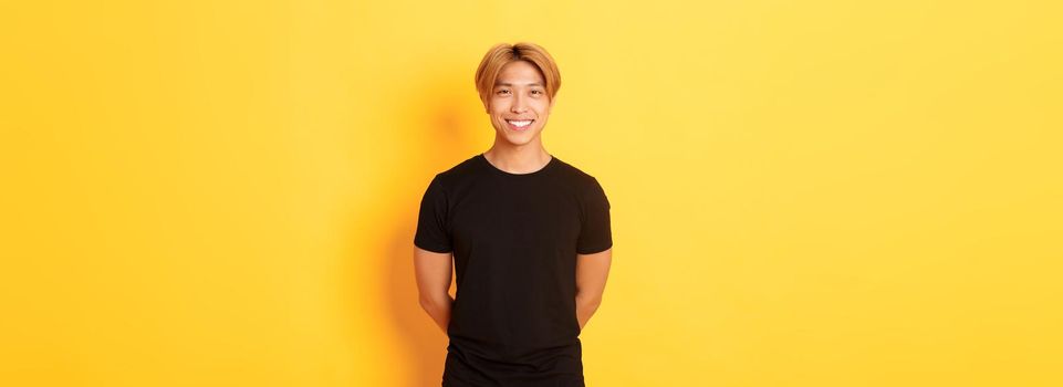 Portrait of friendly handsome asian guy with blond hair, smiling politely, holding hands behind back, standing yellow background.