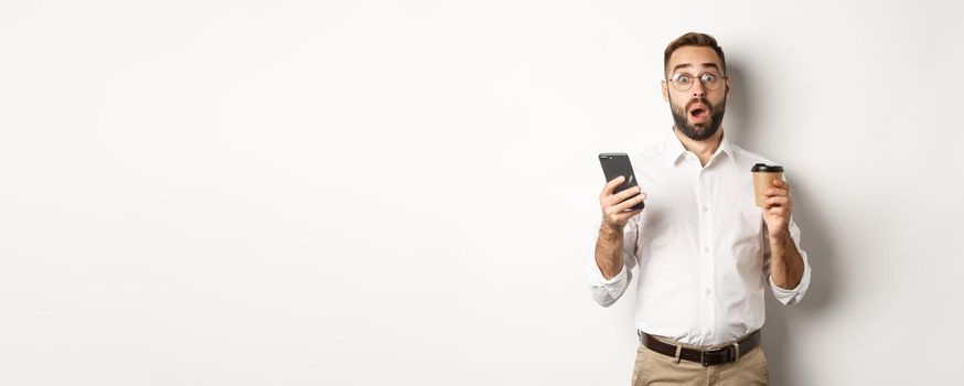 Image of handsome manage drinking coffee, reacting surprised to message on mobile phone, standing over white background.