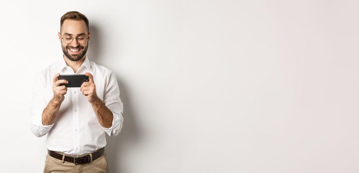 Happy businessman watching video on mobile phone, standing over white background.