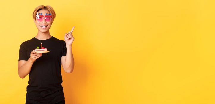 Cheerful, handsome asian birthday guy in funny sunglasses, holding b-day cake with candle and pointing finger upper right corner at your logo, yellow background.