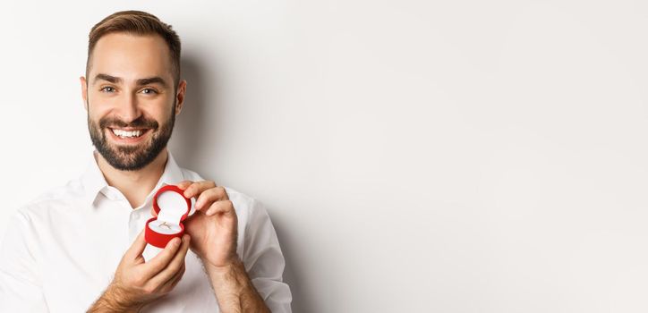 Close-up of happy handsome man making a proposal, holding wedding ring in box and smiling, asking to marry him, white background.