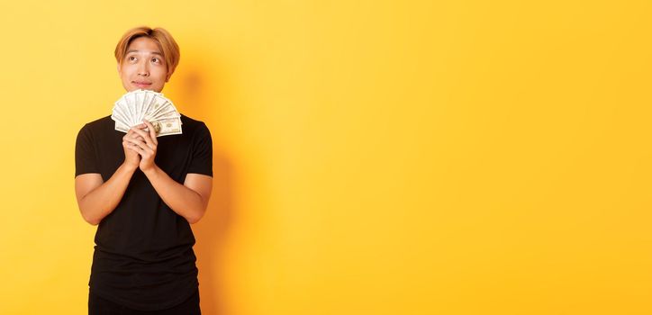 Portrait of dreamy handsome asian guy showing his savings and thinking, looking upper left corner, holding money, standing yellow background.