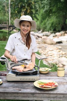 Joyful woman traveller sitting in folding chair and preparing dinner near the river bank. Adventure, travel and camping concept.