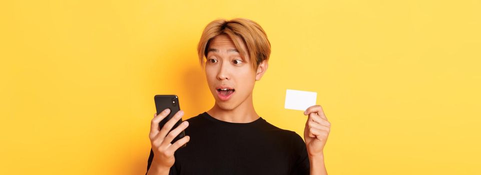 Close-up of amazed asian guy looking at mobile phone while shopping online and showing credit card, yellow background.