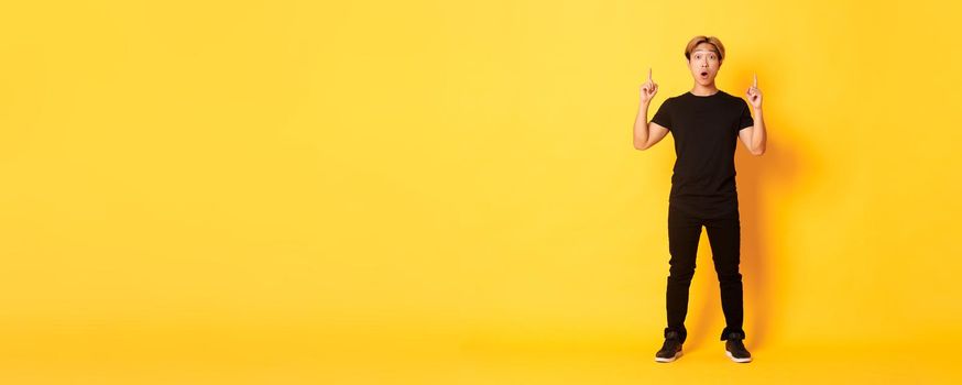 Full-length of impressed asian guy in black clothes, gasping amazed and pointing fingers up, yellow background.