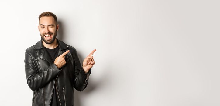 Cheeky guy in leather biker jacket, winking flirty and pointing at upper right corner copy space, white background.