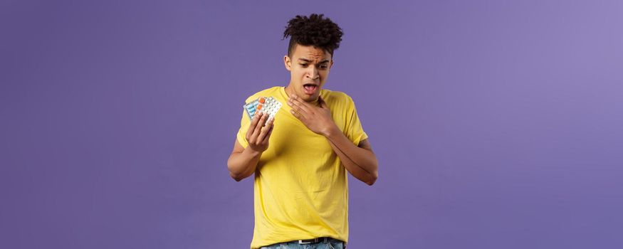 Health, influenza, covid-19 concept. Portrait of young man coughing, choking with tablet, drank pill without water, holding drugs and touch neck as suffer sore throat, purple background.